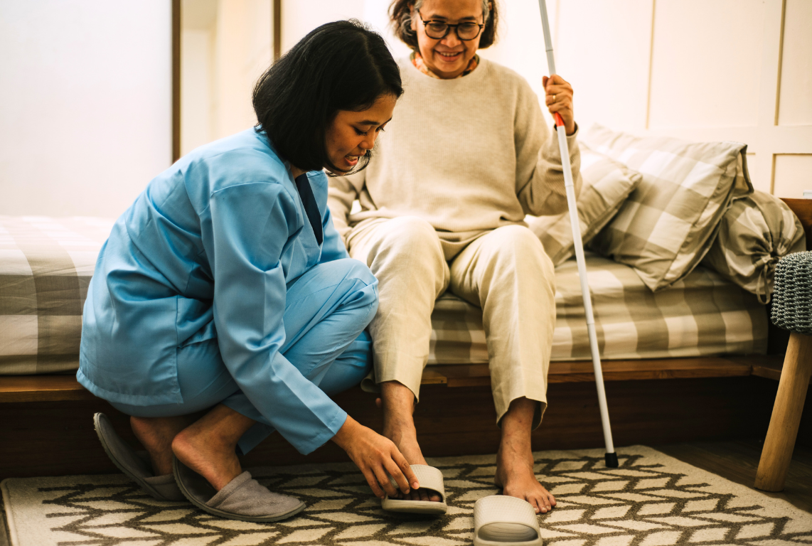 A smiling caregiver kneels beside an elderly woman in a living room, gently helping her put on her shoes.