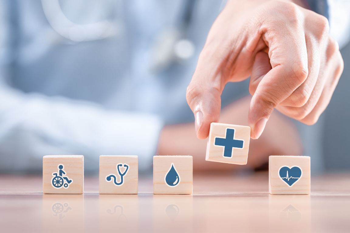 A hand selects wooden blocks stamped with healthcare icons—heart, cross, and calendar—on a tabletop.