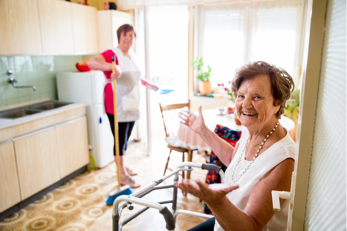 An elderly woman smiles toward the camera while a caregiver sweeps the floor behind her in a sunlit room.