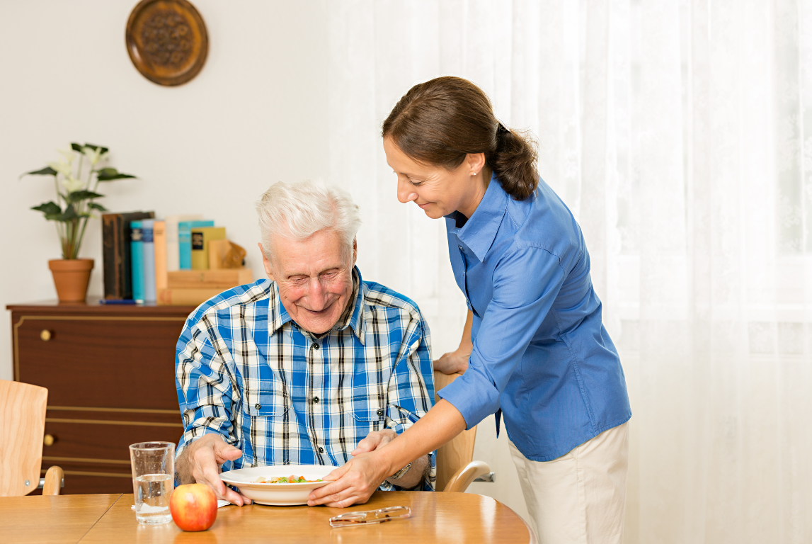 A senior man reaches for a plate of fresh fruit and water served by a caregiver at a dining table