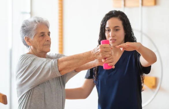 Supportive female caregiver guiding elderly woman through arm-strengthening exercises with hand weights in a bright home setting