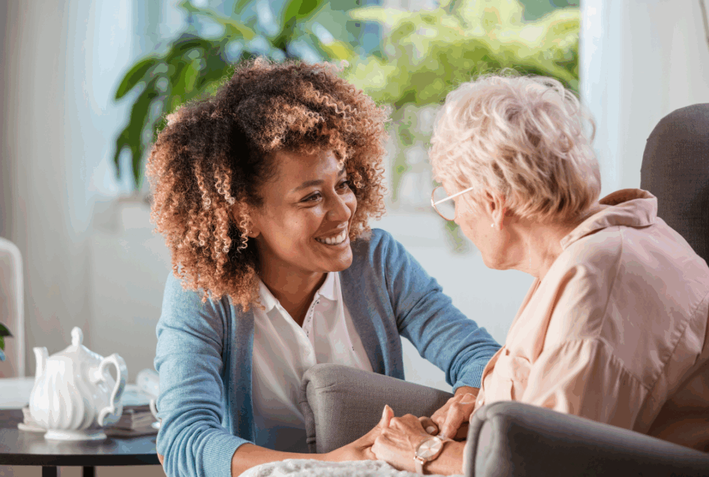 A kind caregiver sits beside a senior in a comfortable chair, warmly holding the senior’s hands in a living room.