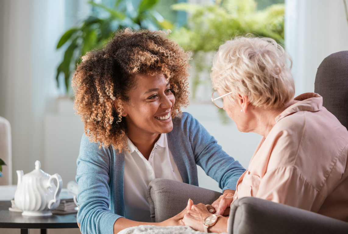 A kind caregiver sits beside a senior in a comfortable chair, warmly holding the senior’s hands in a living room.